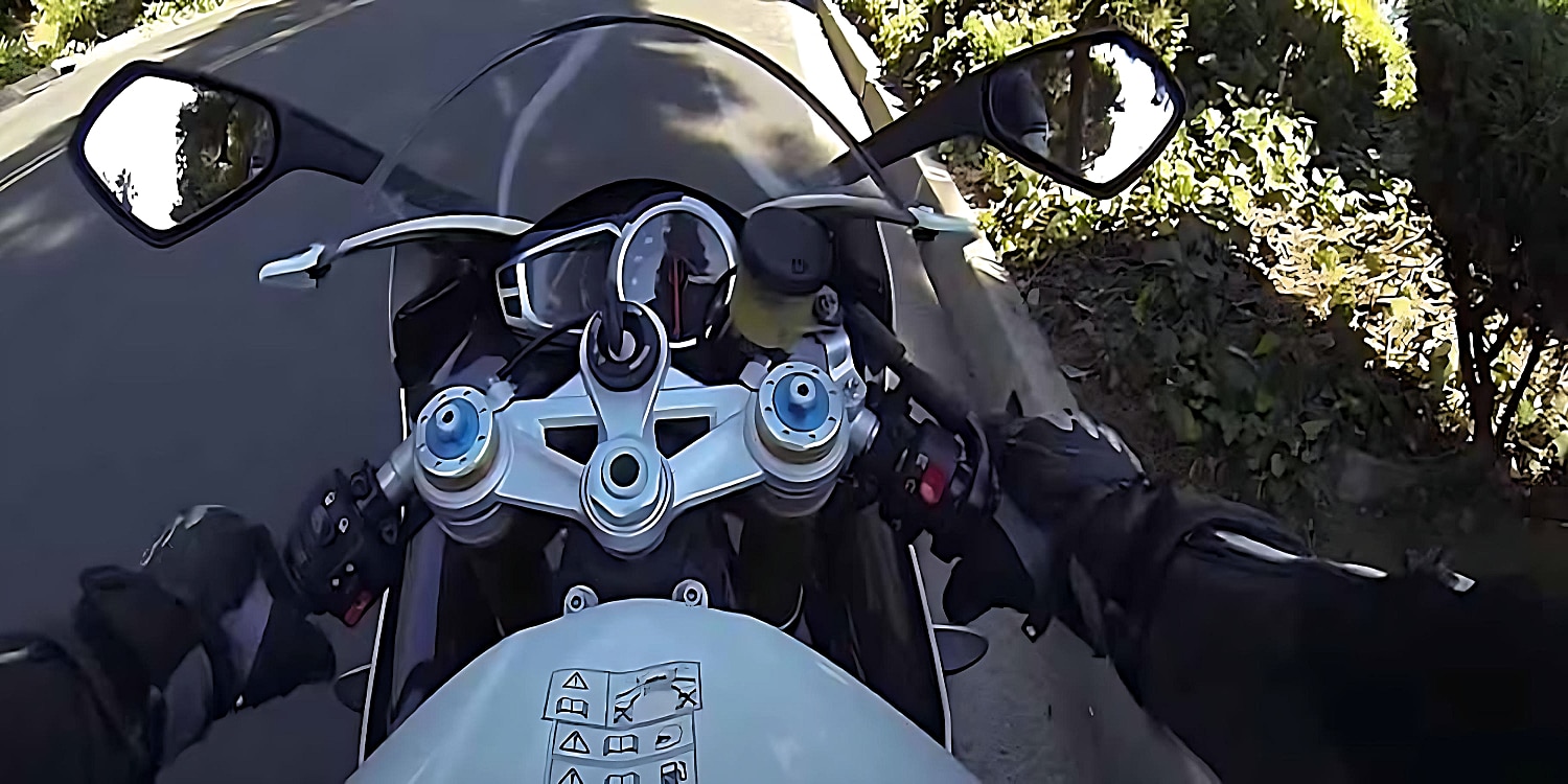 A rider on a sport motorcycle prepares to take off on a pavement surrounded by greenery, with their speedometer still showing 0 mph.