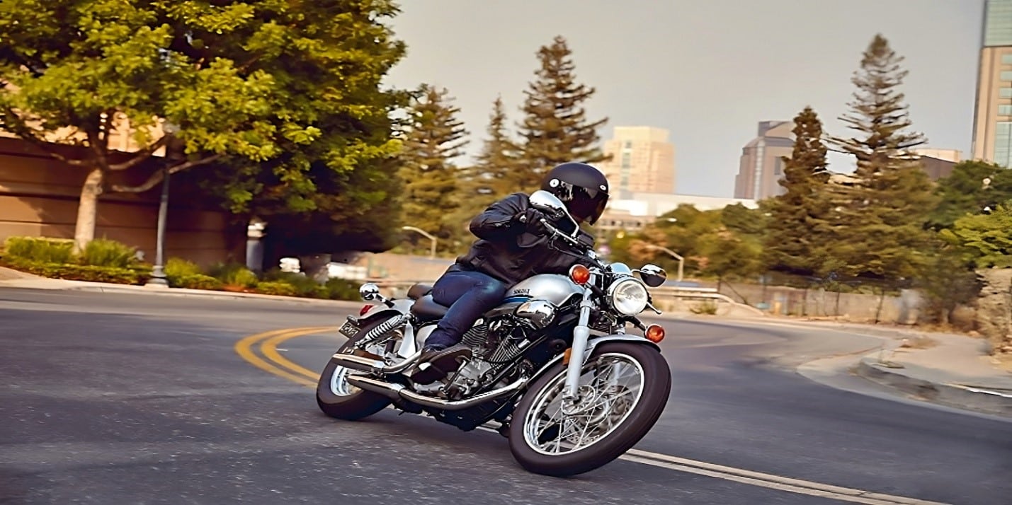 A silver Yamaha V Star 250 motorcycle leaning into a corner on a winding road. The rider is bent low, showcasing the bike's agile handling and responsiveness. The 250cc engine, comfortable seating, and smooth ride make this motorcycle a top pick for both experienced and novice riders
