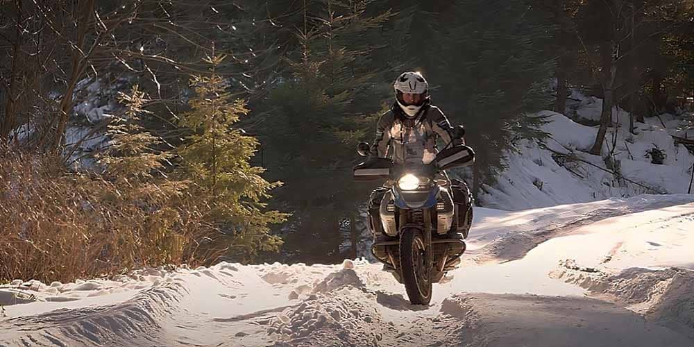 A motorcycle rider cruising down a snowy rural road on a BMW 1100 GS with tall trees in the background, braving the cold weather for an adventurous ride. Cold weather motorcycle riding can be enjoyable, but it also carries some risks that you should be aware of and take necessary precautions.