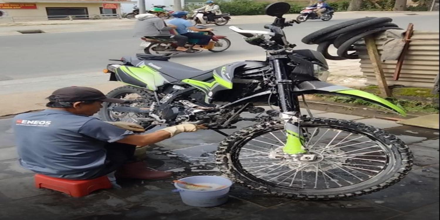 Vietnam: A man donning protective washing gloves scrubs the bash plate of a Kawasaki KLX 250 dual-purpose motorcycle by the roadside. In the background, other riders go about their business.