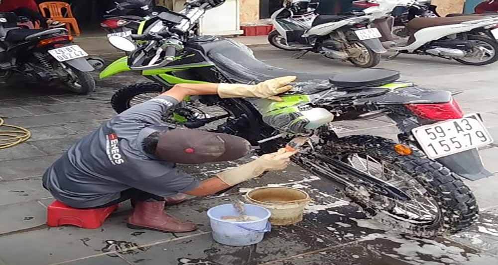 Seated on a short stool to access the underside of the motorcycle. Motorcycle chains come with factory-installed grease on the inside sealed from the exterior elements by rubber gaskets. These gaskets are susceptible to high-pressure water jets and can leak, losing chain lubrication, efficiency, and longevity.