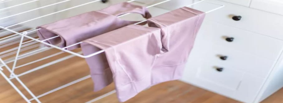 A set of pink of motorcycle base layer hanging from a sturdy and spacious white drying rack, airing out to dry.