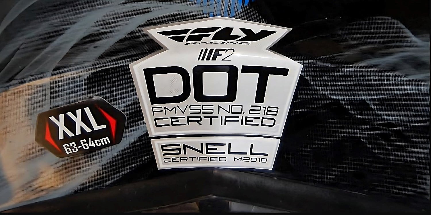A motorcycle helmet displaying both the DOT FMVSS No.218 sticker and the SNELL M2020 sticker, indicating its compliance with both standards. The helmet size is XXL (63-64cm)