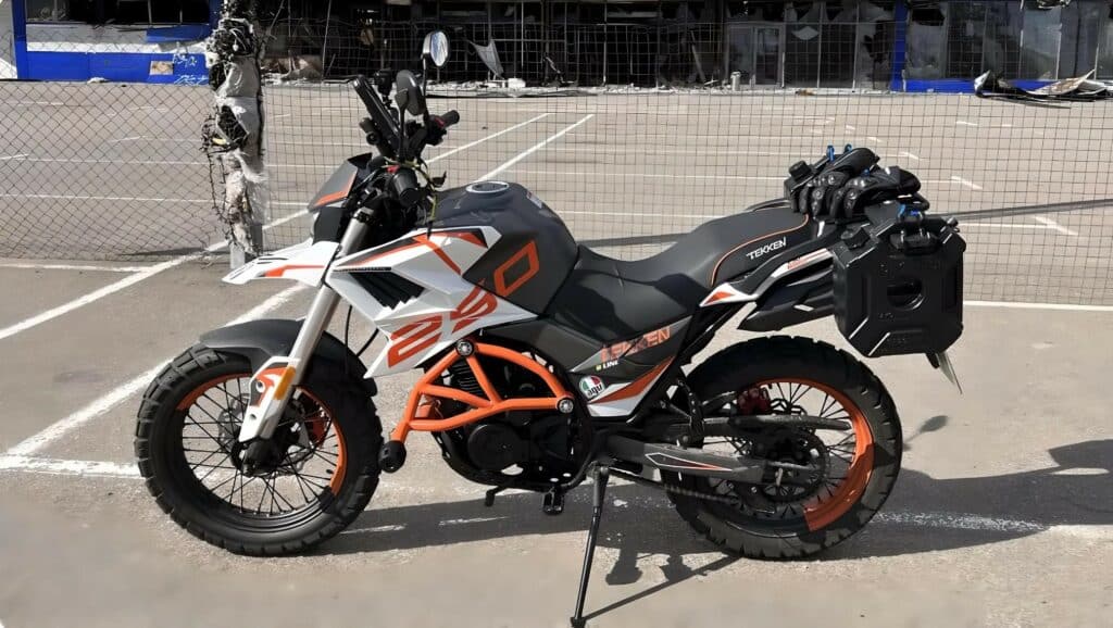 The Tekken 250 adventure touring motorcycle stands in front of the Epicentr in Bucha, Ukraine, with the dual auxiliary tanks mounted at the back. I had initially installed them on the Kawasaki KLX 250 and later transferred them to the Tekken. These tanks can be fitted onto any dual sport, enduro, or adventure touring bike; however, they are not suitable for cafe racers. The only requirement for a motorcycle to utilize these tanks is to have the small metal "rack" positioned behind the passenger seat.